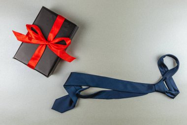 Şık bir mavi kravat kırmızı kurdeleyle süslenmiş güzel paketlenmiş bir hediye kutusunun yanında yatıyor. Bu kurulum, Babalar Günü ve doğum günü kutlamaları için düşünceli hediye seçeneklerinin altını çiziyor..