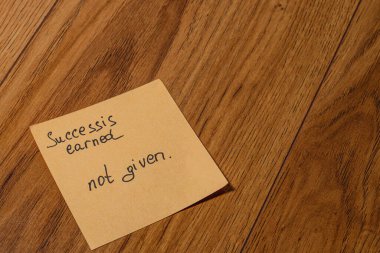 Elle yazılmış bir not ahşap zeminde duruyor, başarının kazanıldığı ve basitçe verilmediği hakkında güçlü bir mesaj iletiyor, ilham verici bir yansıma ve motivasyon..