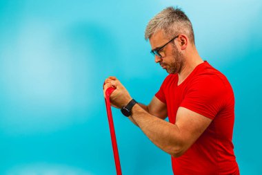 Kırmızı gömlekli ve gözlüklü bir adam dayanıklılık eğitimi için direnç bandını kullanmaya odaklanıyor. Katı mavi arka plan spor için canlı bir ortam yaratıyor..