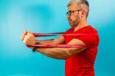 Kırmızı gömlekli bir adam evde direniş bandı egzersizleri yapıyor. Formuna odaklanıyor, açık mavi bir arka plana karşı güç gösterisi yapıyor..