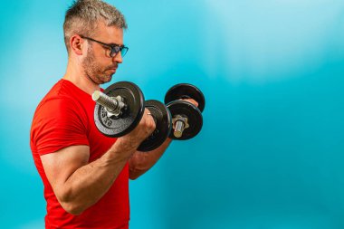 Kırmızı gömlekli bir adam parlak mavi arka planda halterli biseps yapıyor. Güç toplamaya ve fitness geliştirmeye odaklanıyor..