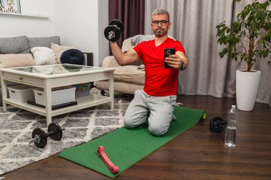 Bir adam oturma odasında yeşil bir egzersiz paspasının üzerine diz çöker, telefonunu tutarken dambılı kaldırır. Spor aletleri onu çevrelemiş, aktif bir yaşam tarzı olduğunu gösteriyor..