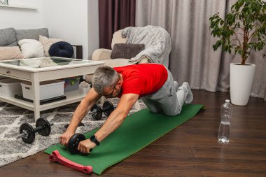 Kırmızı gömlekli bir adam, oturma odasındaki yeşil bir paspasın üzerinde karın kasları egzersizi yapıyor. Dambıllar ve su şişesi yakınlarda, antrenman düzenini vurguluyorlar..