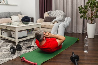 Bir adam, rahat bir oturma odasında yeşil bir paspasın üzerinde karın egzersizleri yapıyor. Mekanda bir kanepe, bitkiler ve spor aletleri var. Ev ortamında bir spor salonu ortamı yaratıyor..