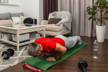 Kırmızı tişörtlü yaşlı bir adam oturma odasındaki yeşil bir paspasın üzerinde şınav çeker. Egzersiz ekipmanları ve rahatlatıcı mobilyalarla çevrilidir..