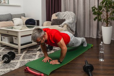 Bir adam, rahat bir oturma odasında yeşil bir spor minderinin üzerinde şınav çekiyor. Dambıllar ve su şişesi yakınlarda. Bu da rutin bir egzersiz olduğunu gösteriyor..