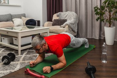 Bir adam şık bir oturma odasında yeşil bir paspas üzerinde tahta egzersizi yapıyor. Dekorun yanında rahat bir kanepe, ağırlık ve su torbası var..