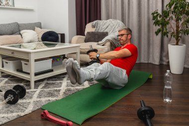 Kırmızı gömlekli bir adam, ağırlıklar ve su şişesiyle çevrili yeşil bir yoga minderinin üzerinde karın egzersizi yapıyor..