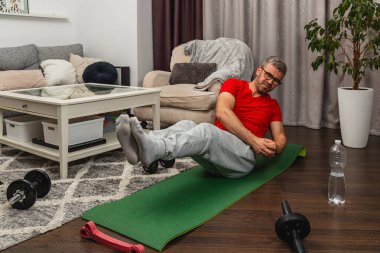 Kırmızı gömlekli ve gri eşofmanlı bir adam yeşil bir paspasın üzerinde, bitki ve mobilyalarla süslenmiş bir oturma odasında egzersiz yapıyor..