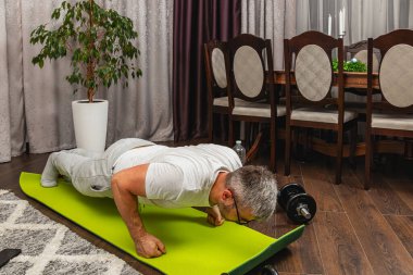 Formda bir adam, sıcak oturma odasında parlak yeşil bir spor minderinin üzerinde şınav çekiyor. Sofra bir yemek masası ve saksı bitkisi içeriyor. Bu da evde aktif bir yaşam tarzı olduğunu gösteriyor..