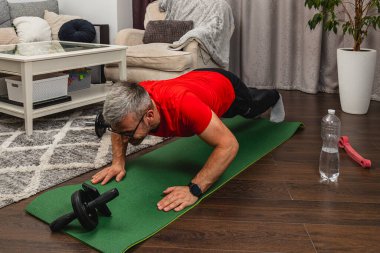 Kırmızı tişörtlü bir adam, rahat bir oturma odasında yeşil bir paspasın üzerinde şınav çekiyor. Yakınlarda bir su şişesi ve ağırlıklar var, evde çalışma ortamı yaratıyor..