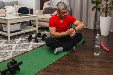 Kırmızı tişörtlü bir adam oturma odasındaki yeşil yoga minderinde bacak bacak üstüne atmış bir şekilde oturuyor. Yanında halterler ile antrenman yapmaya hazırlanırken takip cihazını izliyor..