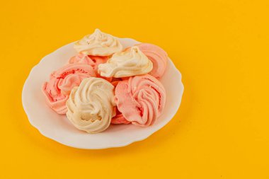 Bir tabak çeşitli pembe ve beyaz kremalı kurabiyeler içerir, yumuşak dokularını sergiler ve parlak sarı bir arka plana karşı kıvrılmış tasarımlar sergilerler..