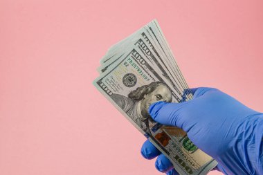 Eldivenli bir el birkaç yüz dolarlık banknotları yumuşak pembe bir fon üzerinde tutar, para ve koruyucu ekipman arasındaki kontrastı vurgulayarak.