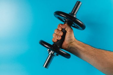 Bir el gümüş uçlu siyah bir dambılı kavrıyor, güç ve odaklanma gösteriyor. Parlak mavi arkaplan etkinliği ve kararlılığı vurguluyor.