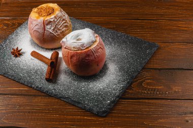 İki fırınlanmış elma servis tahtasının üzerinde pudra şekeriyle tozlanmış şekilde otururlar. Tarçın çubukları ve yıldız anason onlara eşlik ederek sıcak ve davetkar bir ortam oluşturur..