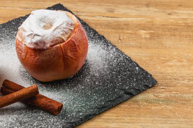 Pudra şekerli fırınlanmış bir elma koyu bir tahta üzerinde oturur. Tarçın çubukları yakına yerleştirilir, sonbahar için mükemmel sıcak bir atmosfer yaratır..