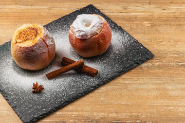 Pudra şekerli iki fırınlanmış elma koyu bir tepside oturur. Yakınlarda, iki tarçın çubuğu ve bir yıldız anasonu sıcacık sonbahar atmosferine eklenir.