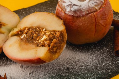 Karamel karışımı ile dolu fırınlanmış elmalar koyu bir levhada, pudra şekeriyle serpilmiş, ve tarçın çubuklarıyla çevrili, rahat bir atmosfer yaratıyor..