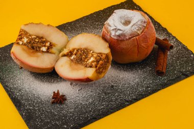 İki fırınlanmış elma siyah bir levhada sergilenir. Bir elma, fındık ve baharattan yapılmış lezzetli bir dolguyu ortaya çıkarmak için ikiye bölünür. Parlak sarı arka plan renkleri güçlendirir.