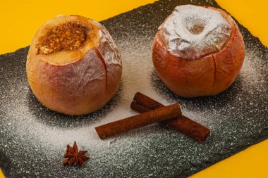 Pudra şekerli ve tarçın çubuklarıyla çevrili iki fırınlanmış elma ve bir yıldız anason parlak sarı bir zemin üzerinde siyah bir tahta tepside oturur..