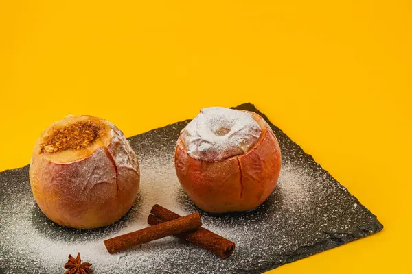 İki fırınlanmış elma pudra şekerle kaplanmış ve tahta tepside tarçın çubuklarıyla servis edilmiş, parlak sarı bir arkaplana karşı sıcak ve davetkar bir tatlı gösterisi yaratıyor..