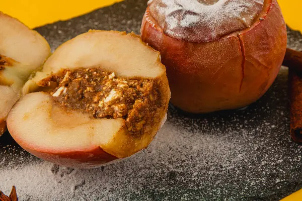 Karamel karışımı ile dolu fırınlanmış elmalar koyu bir levhada, pudra şekeriyle serpilmiş, ve tarçın çubuklarıyla çevrili, rahat bir atmosfer yaratıyor..