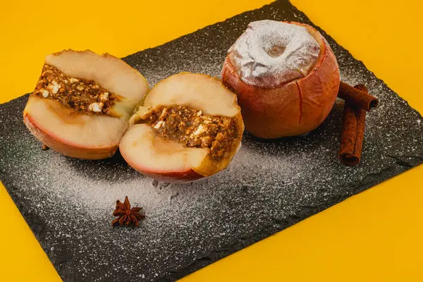 İki fırınlanmış elma siyah bir levhada sergilenir. Bir elma, fındık ve baharattan yapılmış lezzetli bir dolguyu ortaya çıkarmak için ikiye bölünür. Parlak sarı arka plan renkleri güçlendirir.