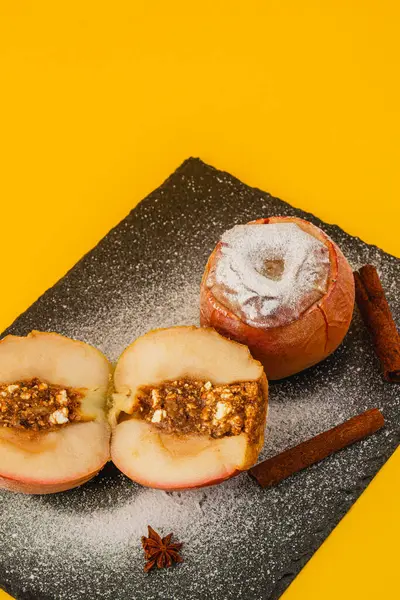 İki fırınlanmış elma karanlık bir tahta yüzeyde oturur. Bir elma ikiye bölünür, içinde tatlı bir dolgu ortaya çıkar, yanına tarçın çubukları yerleştirilir..