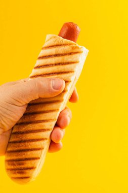 Bir el, içinde sosisli sandviç olan tost ekmeğini parlak sarı bir arka planda kavrayıp her durum için mükemmel bir aperatif sunuyor..