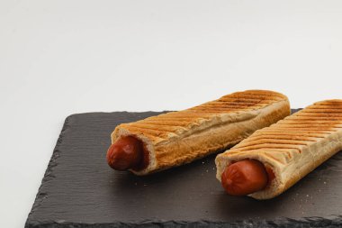 Kızarmış ekmeklere konmuş iki ızgara sosisli, siyah bir tabakta dinleniyor. Basit, hızlı ve doyurucu bir atıştırmalık ya da yemek için idealdir..