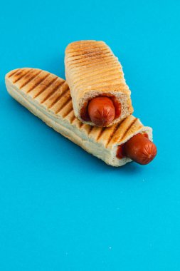 Kızarmış ekmeğin içine yerleştirilmiş iki ızgara sosisli sandviç parlak mavi bir arka planda sergileniyor. Altın kabuk, bu atıştırmalığın görsel çekiciliğini arttırır..