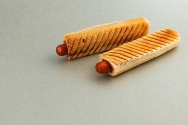 İki ızgara sosisli sandviç, çapraz ızgara izleriyle sıcak, kızarmış ekmeğin içine yerleştirilir. Yemek tarafsız bir arkaplanda sunuluyor, dokusunu vurguluyor..