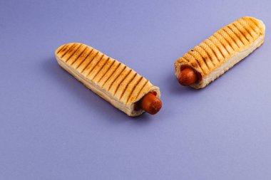 Kızarmış çöreğin içine iki ızgara sosisli yerleştiriliyor. Katı mavi arka planda lezzetli bir yemek sergileniyor. Çöreklerde ilave doku ve cazibe için ızgara izleri var..