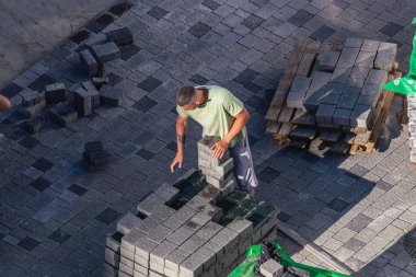 Lviv Ukrayna 04.30.25 Bir işçi inşaat alanında gri kaldırım taşlarını dikkatle dizer. Sitede organize ahşap paletler ve arka planda tuğlalarla dolu düz bir yüzey yer alıyor..