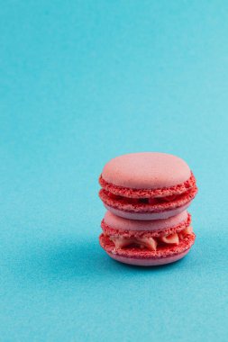 Açık krema dolgulu üç pembe makarna üst üste istiflenmiş, parlak mavi bir zemin üzerine kurulmuş, görsel olarak çekici bir tatlı gösterisi yaratıyor..