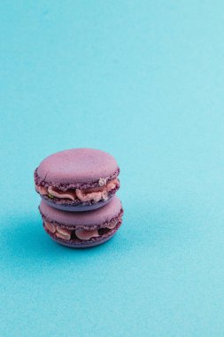 Çikolata kremalı iki mor badem kurabiyesi düz mavi bir zemine karşı düzgünce istiflenmiştir. Narin dokularını ve canlı renklerini sergilerler..