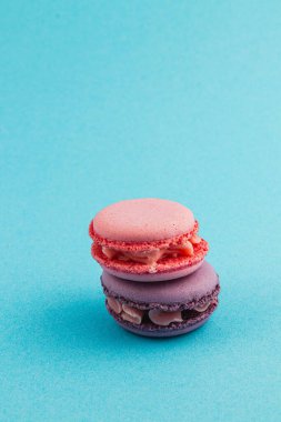 İki badem kurabiyesi, biri pembe, biri mor, parlak mavi bir yüzeye dikkatlice istiflenmiş, pürüzsüz dokularını sergiliyorlar ve enfes dolgularını, davetkar tatlılıklarını sergiliyorlar..