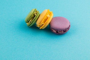 Yeşil, sarı ve mor renkli üç canlı badem kurabiyesi parlak mavi bir yüzeye oturmuş, eşsiz renklerini ve dokularını sergiliyorlar, tatlı bir ziyafeti davet ediyorlar..