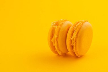 İki sarı badem kurabiyesi, parlak sarı bir yüzeyde yan yana otururlar. Yumuşak dokularını ve ılık ışık altında kremsi dolgularını sergilerler..