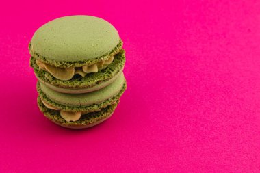 Kremsi dolguyla doldurulmuş üç yeşil badem kurabiyesi parlak pembe bir yüzeye güzelce istiflenmiş, dokularını ve renk kontrastlarını vurguluyorlar..