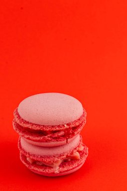 İki pembe badem kurabiyesi birbiri üzerine yığılmış canlı kırmızı bir zemin üzerinde, desenli kabuklarını ve kremsi dolgularını vurguluyorlar..