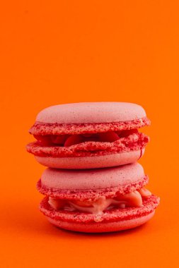 Kremayla dolu iki katman pembe badem kurabiyesi çarpıcı bir turuncu zemine karşı dimdik ayakta duruyor ve hoş dokularını ve renk kontrastlarını gözler önüne seriyor..