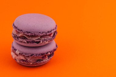 Parlak turuncu bir zemin üzerinde kremalı çikolatalı ganache ile dolu iki istiflenmiş mor badem kurabiyesi renkli ve cezbedici bir tatlı gösterisi yaratıyor..