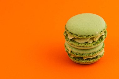 Lezzetli, kremsi dolgulu yeşil badem kurabiyeleri canlı turuncu bir zemin üzerine istiflenerek çarpıcı ve iştah açıcı bir görsel karşıtlık yaratır..