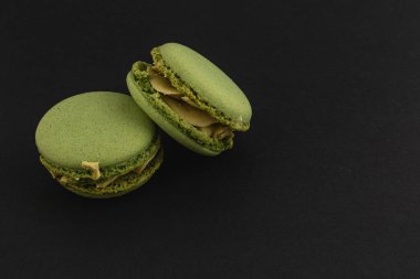 İki canlı yeşil badem kurabiyesi zarif bir şekilde koyu bir zemin üzerinde oturur, narin dokularını ve çekici krema katmanlarını sergilerler..