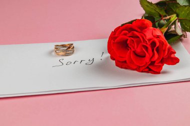 Altın bir nişan yüzüğü, kayıp aşkı ve pişmanlığı sembolize eden canlı kırmızı bir gül eşliğinde, yürekten bir özürle işaretlenmiş beyaz bir zarfın üzerinde duruyor..