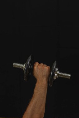 Dambılı kavrayan, güç ve kararlılık gösteren yakın plan bir el. Arka plan karanlık, spor ve ağırlık antrenmanına odaklanmayı vurguluyor..