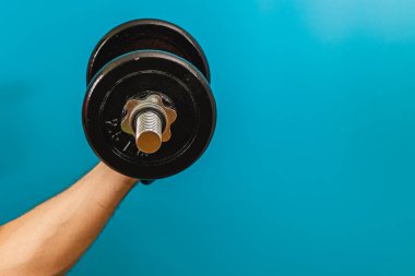 Bir kişi ağır bir dambıl tutuyor ve güç antrenmanı rutinini gösteriyor. Canlı mavi arkaplan spor ve spor üzerine odaklanmayı arttırır.