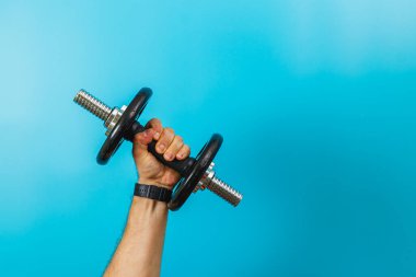 Bir el, parlak mavi arka planda bir halteri sıkıca tutar. Bileği görünür durumda, spor yapan bir fitness meraklısı görünüyor..